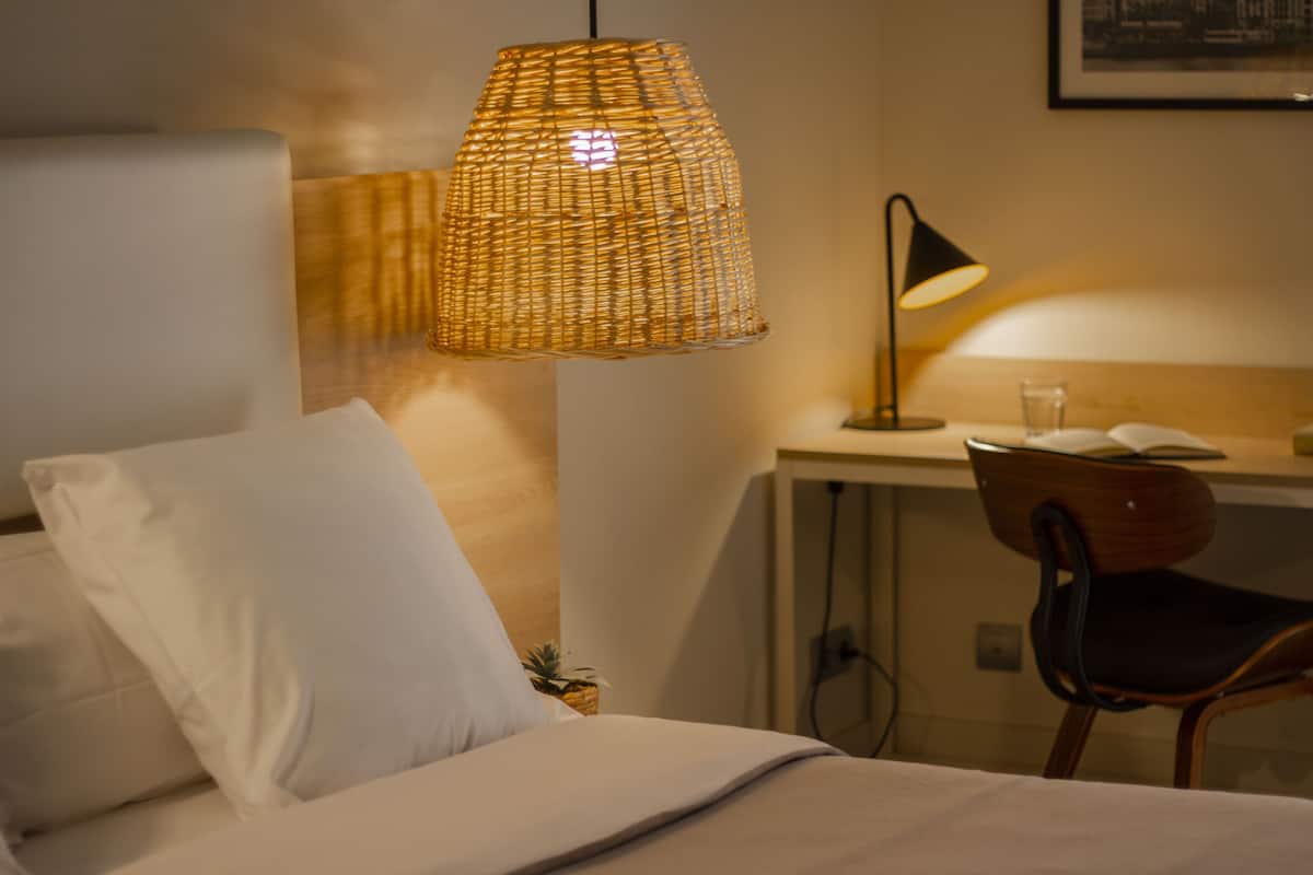 A hotel room with a bed, a desk, a chair, a lamp, and a wall-mounted shelf with a plant.