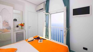 Een hotelkamer met een bed, een klein tafeltje met een bloevas, een raam met uitzicht op zee, en een airconditioner.