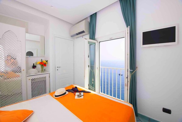 A hotel room with a bed, a small table with a vase of flowers, a window with a view of the sea, and an air conditioning unit.