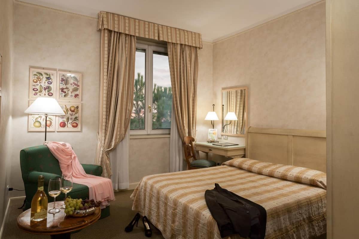 Une chambre d’hôtel avec un lit, un bureau, une chaise, une table avec une bouteille et des verres, et des rideaux.