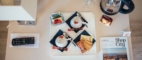 A hotel room table with a remote control, a magazine, a teapot, a cup, a spoon, a glass, a kettle, and some chocolate bars.