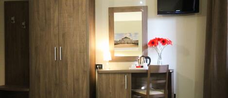 A hotel room with a wooden wardrobe, a desk with a mirror, a chair, and a television on the wall.