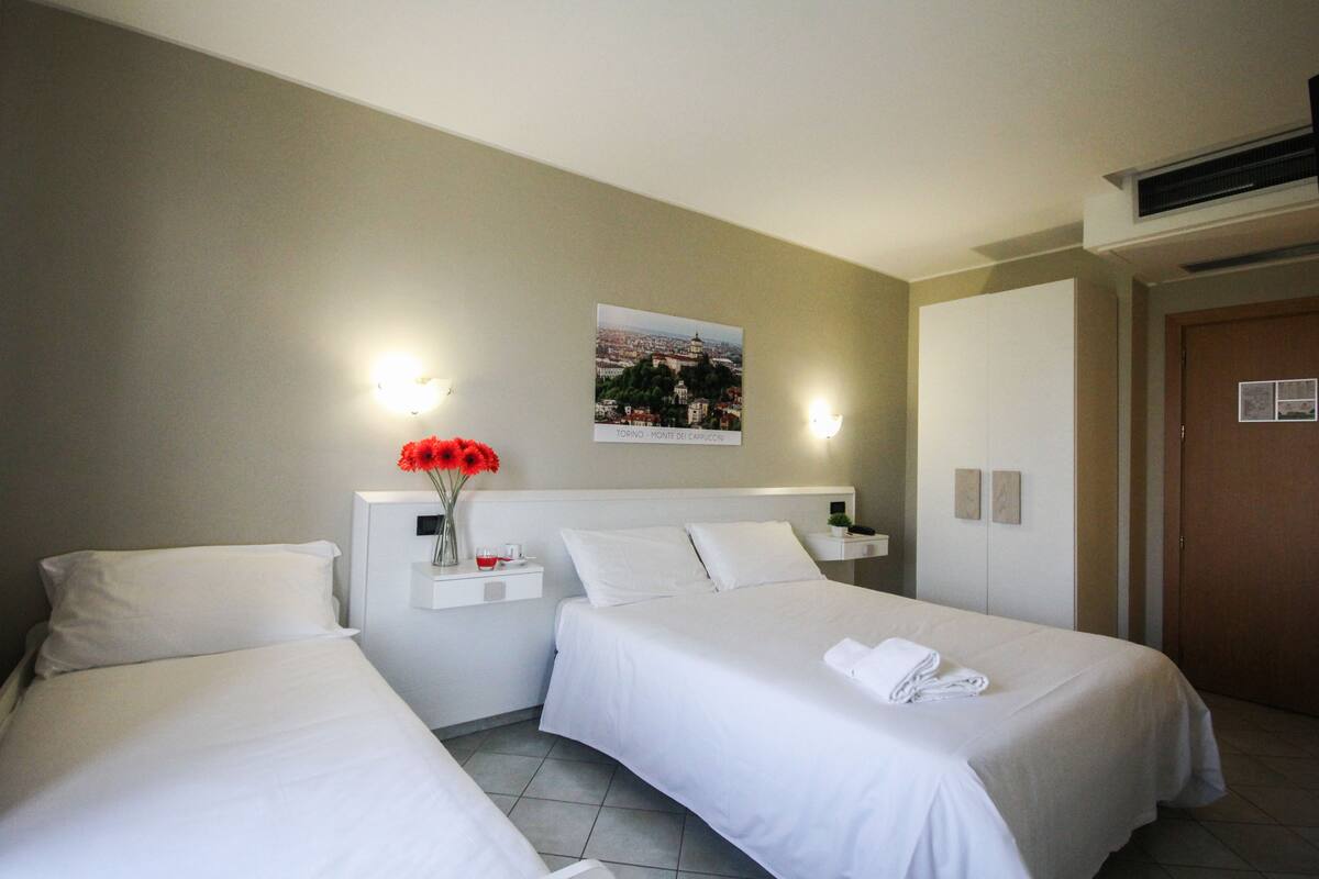 Habitación de hotel con dos camas individuales, una mesita de noche con un florero de flores rojas, un cuadro colgado en la pared y una puerta con interruptor de luz.