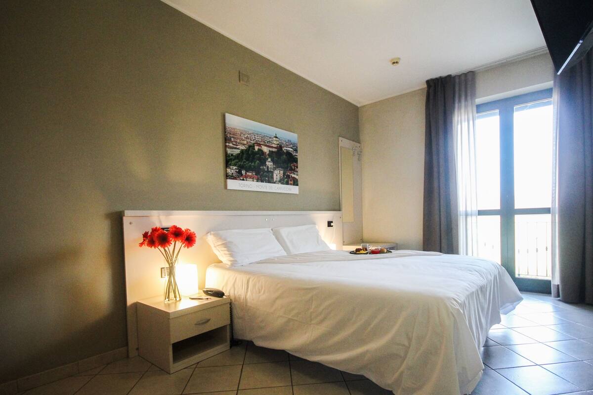 Una habitación de hotel con una cama, una mesita de noche con un florero y un cuadro en la pared, además de una ventana con cortinas.