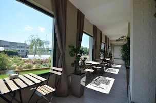 Área externa com mesas, cadeiras, janelas amplas e plantas em vasos.