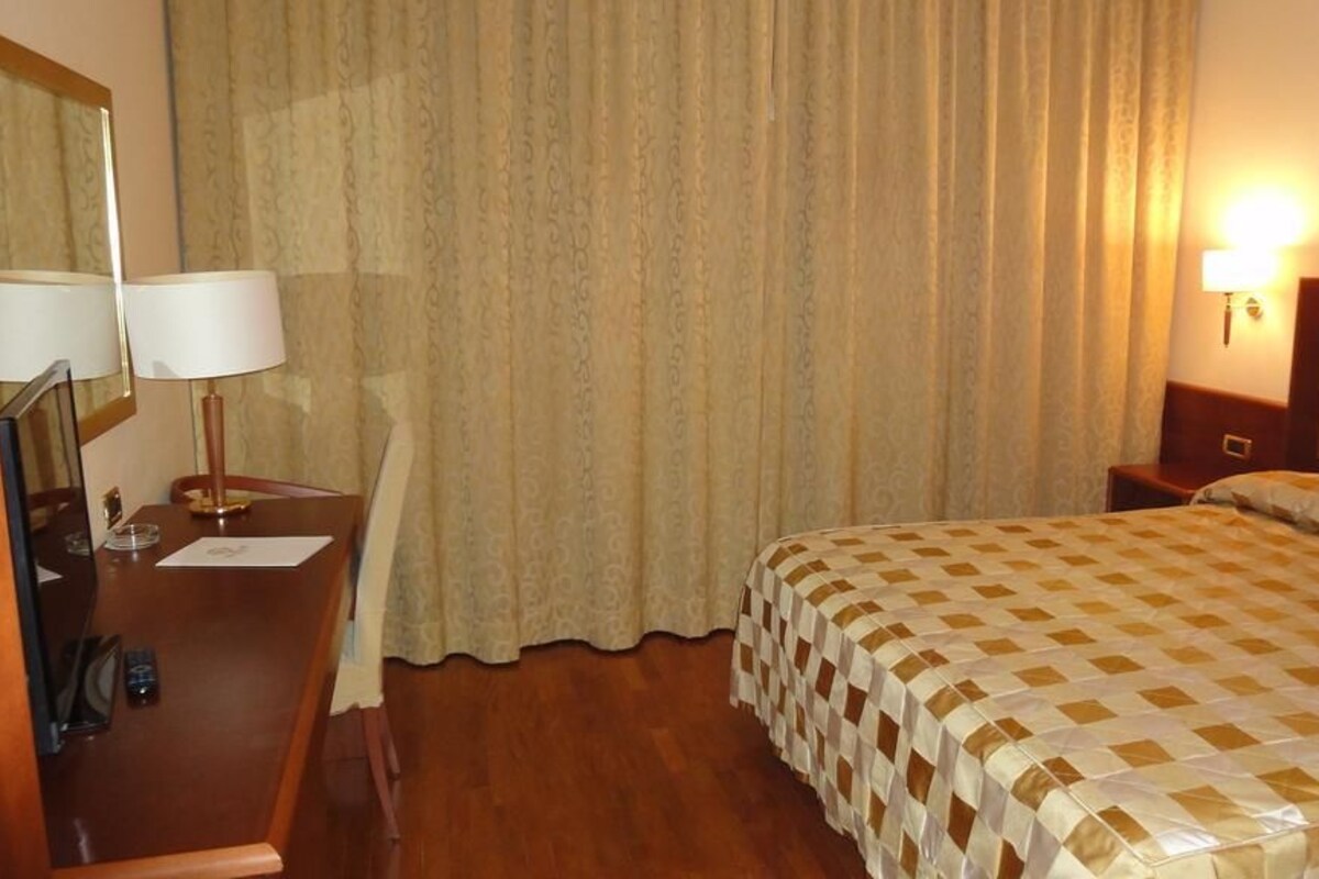 A hotel room with a bed, a desk with a lamp, a mirror, and a wall-mounted light.