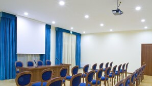Uma sala de conferência com fileiras de cadeiras estofadas em azul, uma mesa de madeira com um microfone, um projetor e uma tela.
