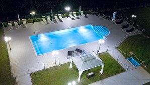 Piscina ao ar livre com espreguiçadeiras e área de estar coberta, cercada por um muro e iluminada por postes de luz.