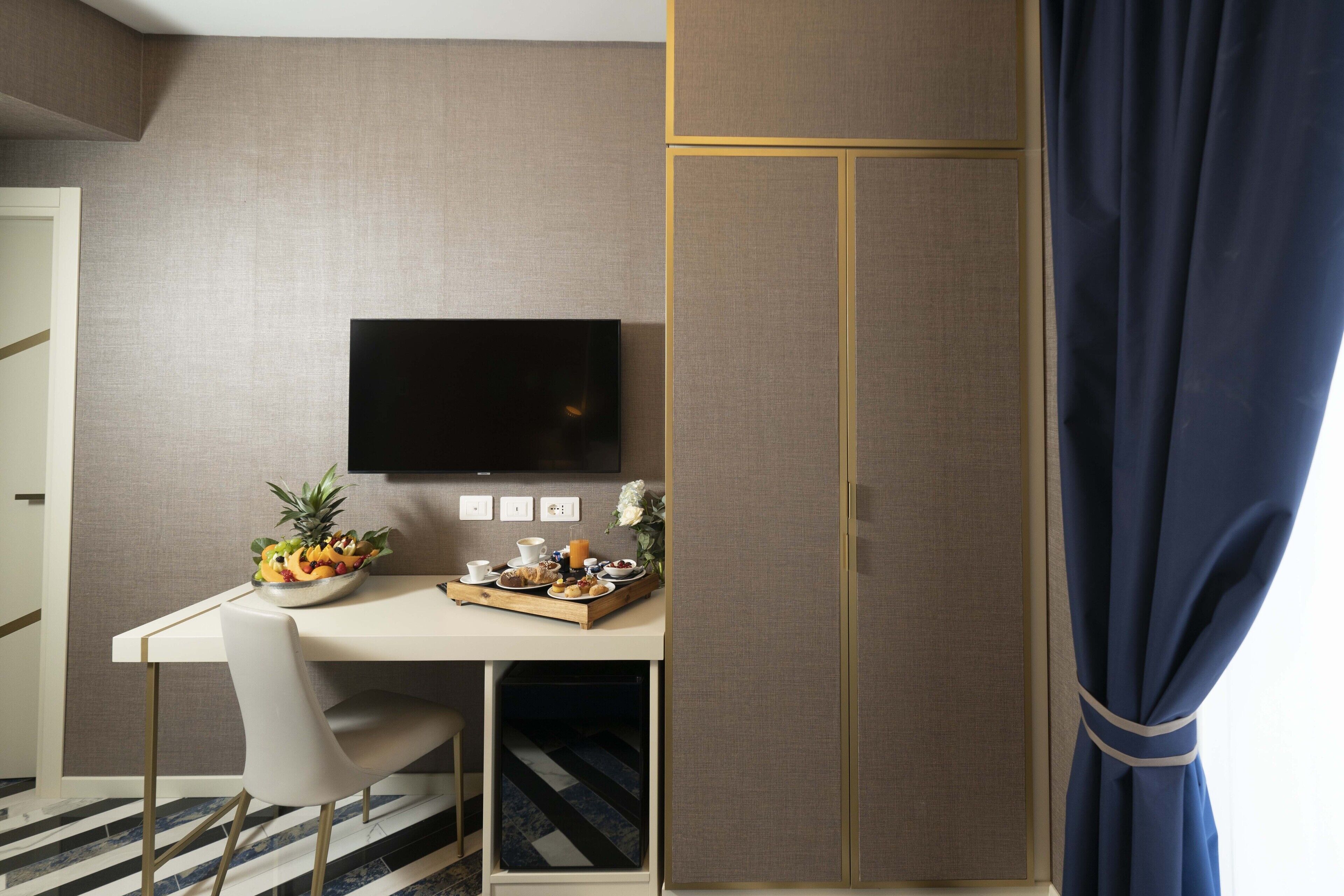 A hotel room with a bed, a desk with a TV, a small table with breakfast items, and a window with curtains.
