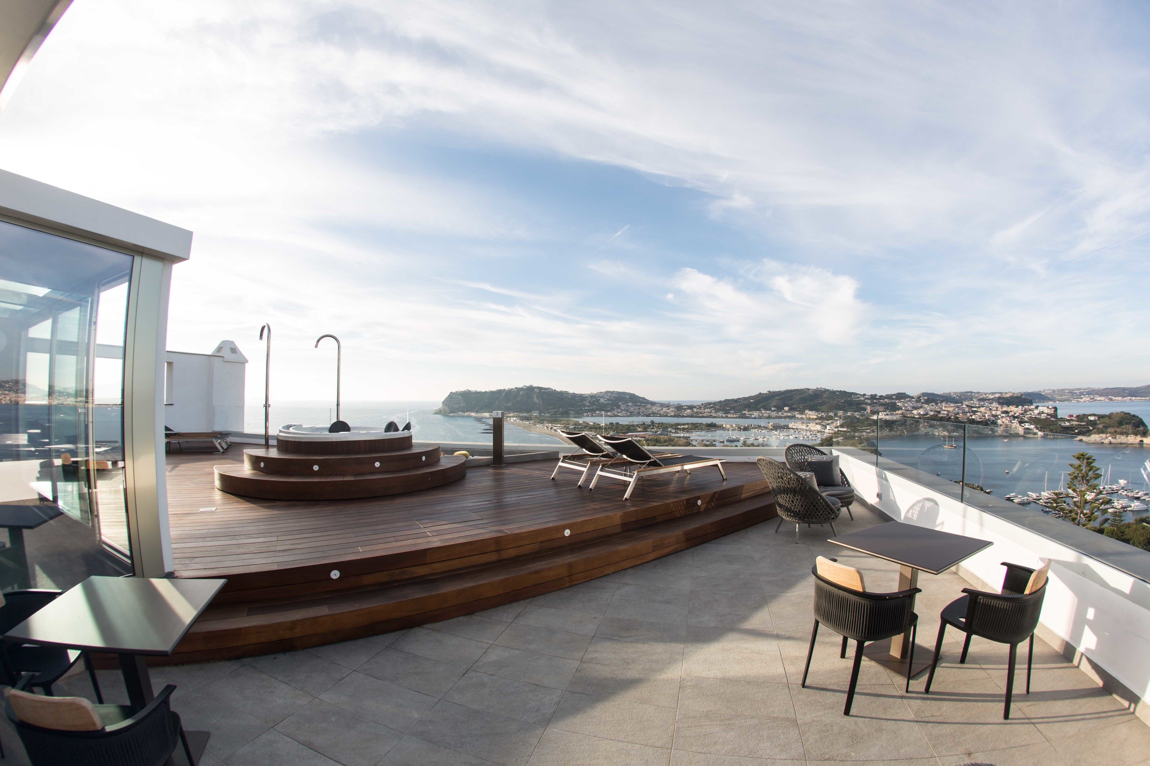 Una terraza en la azotea con cubierta de madera, sillones de descanso y una mesa, con vistas a un pueblo costero y al mar.