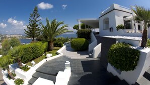 Een modern huis met een witte gevel, een stenen pad dat naar de ingang leidt, en uitzicht op zee.
