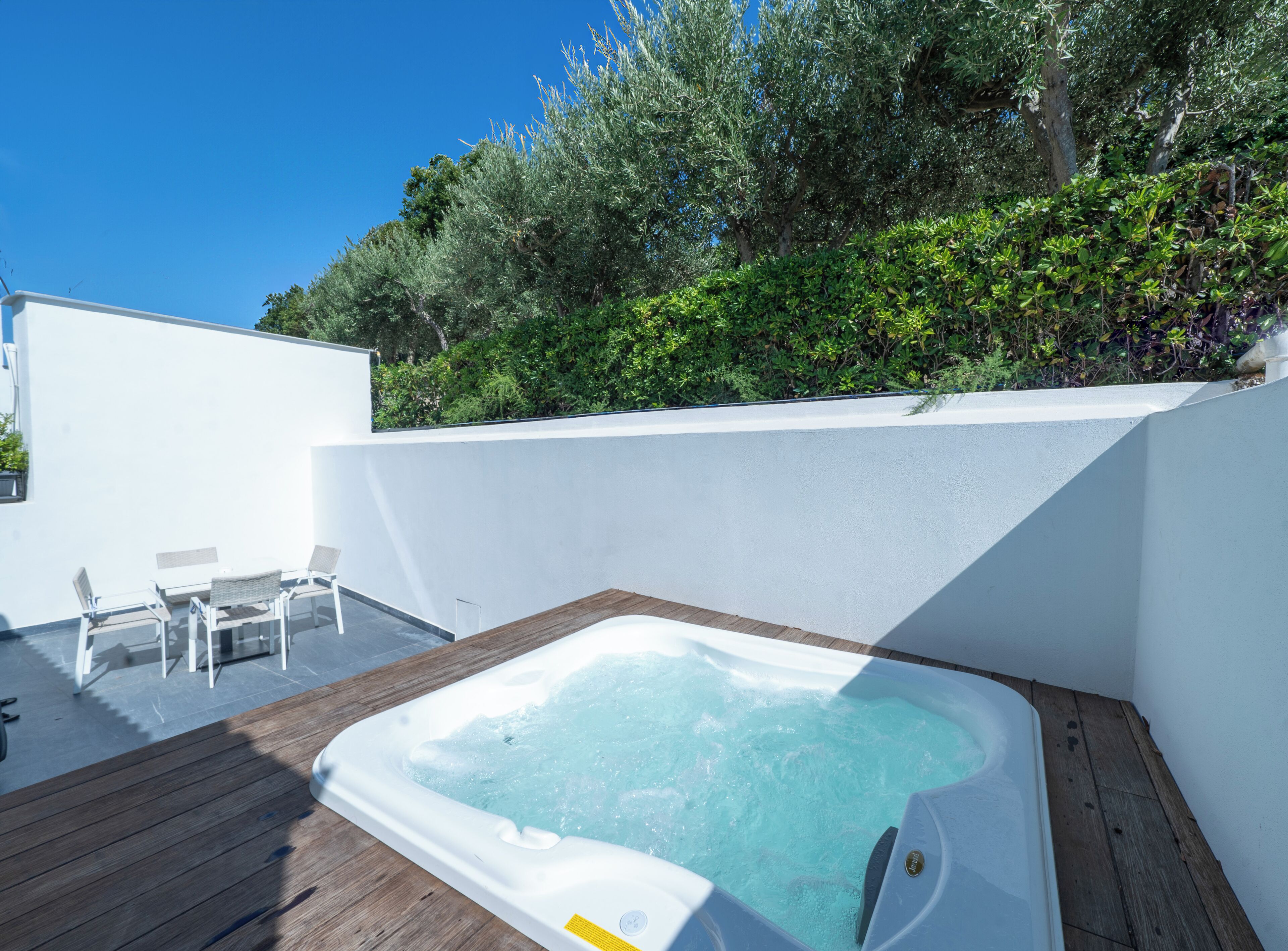 Un bain à remous dans un jardin, avec une terrasse en bois, des murs blancs et une vue sur les arbres et les arbustes.