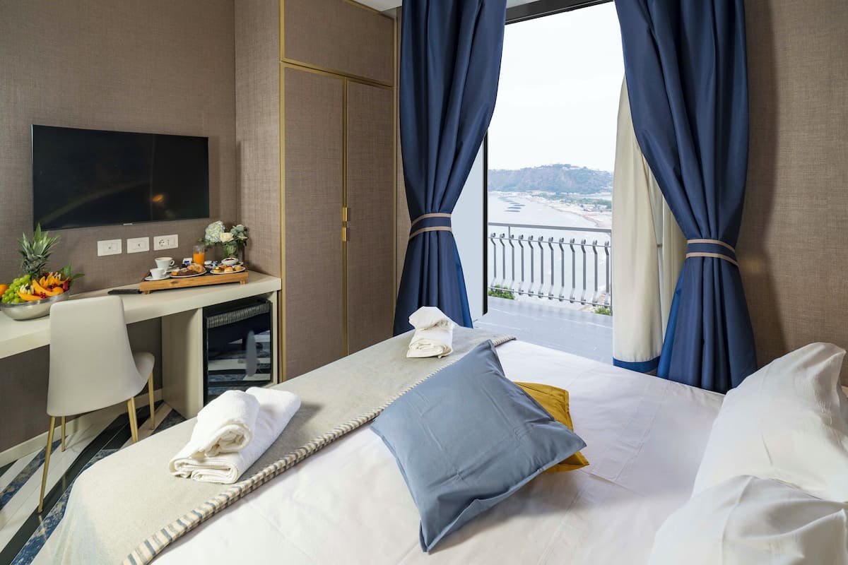 A hotel room with a bed, a desk, a TV, and a view of the sea.