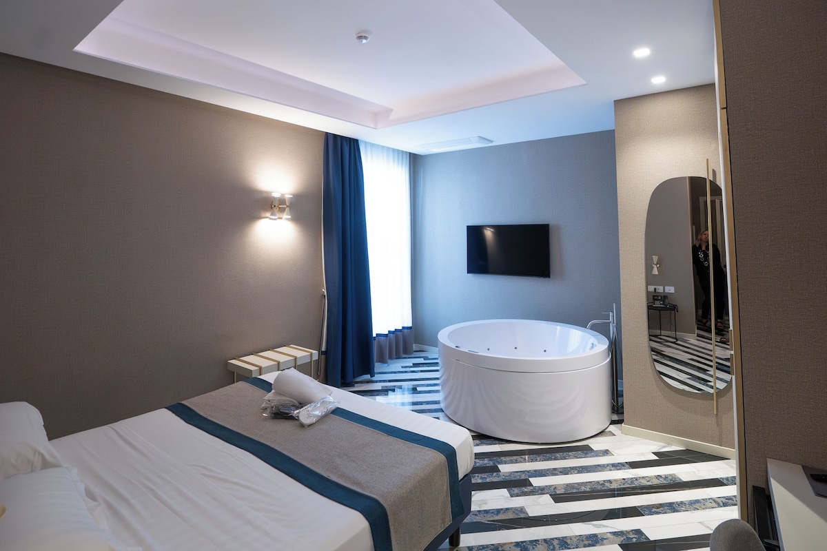 A modern hotel room with a large bed, a separate seating area, a bathtub, and a flat-screen TV.