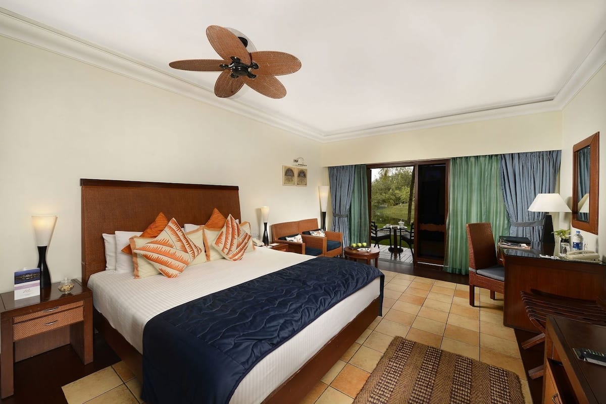 A hotel room with a large bed, a desk, a chair, a ceiling fan, and a view through the glass door.