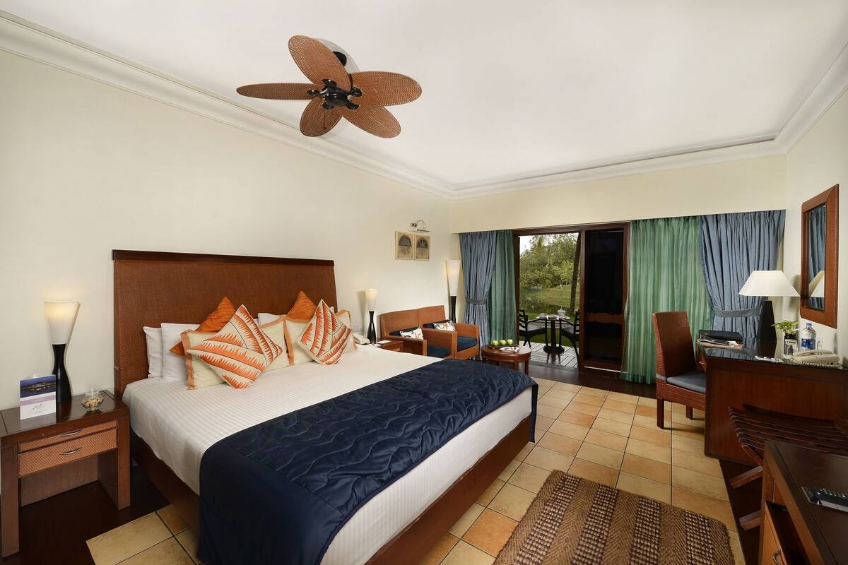 A hotel room with a large bed, a desk with a computer, a chair, a desk lamp, a ceiling fan, and a view of the outdoors.