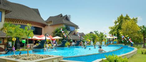 A swimming pool area with a fountain, surrounded by a building with a tiled roof and a covered seating area.