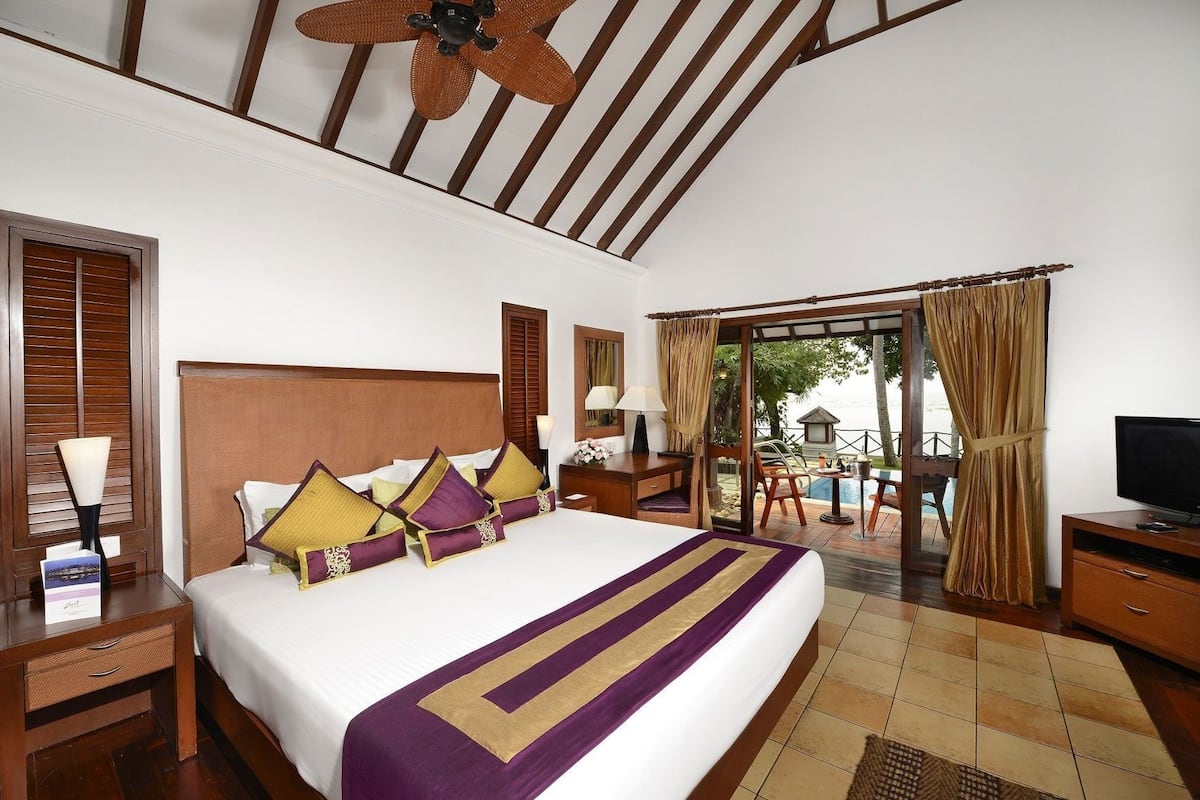 A hotel room with a large bed, wooden headboard, a ceiling fan, a TV, a desk, and a view of the ocean through a glass door.
