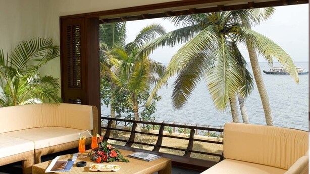 A balcony with wicker furniture, a table with drinks and a menu, and a view of the sea with palm trees and a boat. - The Zuri Kumarakom Kerala Resort & Spa (Kottayam)