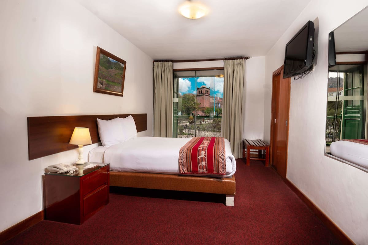 A hotel room with a bed, a bedside table with a lamp, a telephone, a TV mounted on the wall, and a window with curtains.