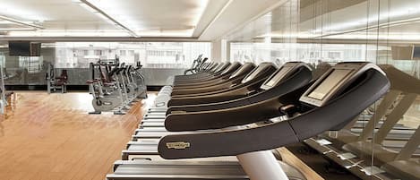 A row of stationary bikes in a gym with a tiled floor and large windows.