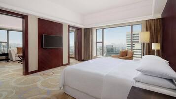 Habitación de hotel con una cama grande, un televisor de pantalla plana en la pared, vistas a la ciudad a través de amplios ventanales, una zona de estar con una silla y una alfombra con diseño circular.