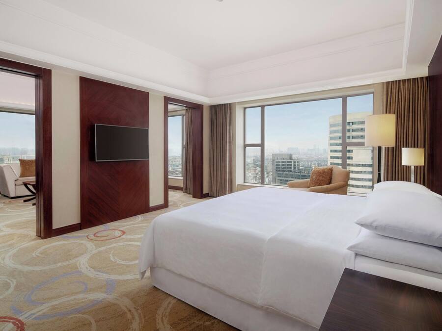 Habitación de hotel con una cama grande, un televisor de pantalla plana en la pared, vistas a la ciudad a través de amplios ventanales, una zona de estar con una silla y una alfombra con diseño circular.