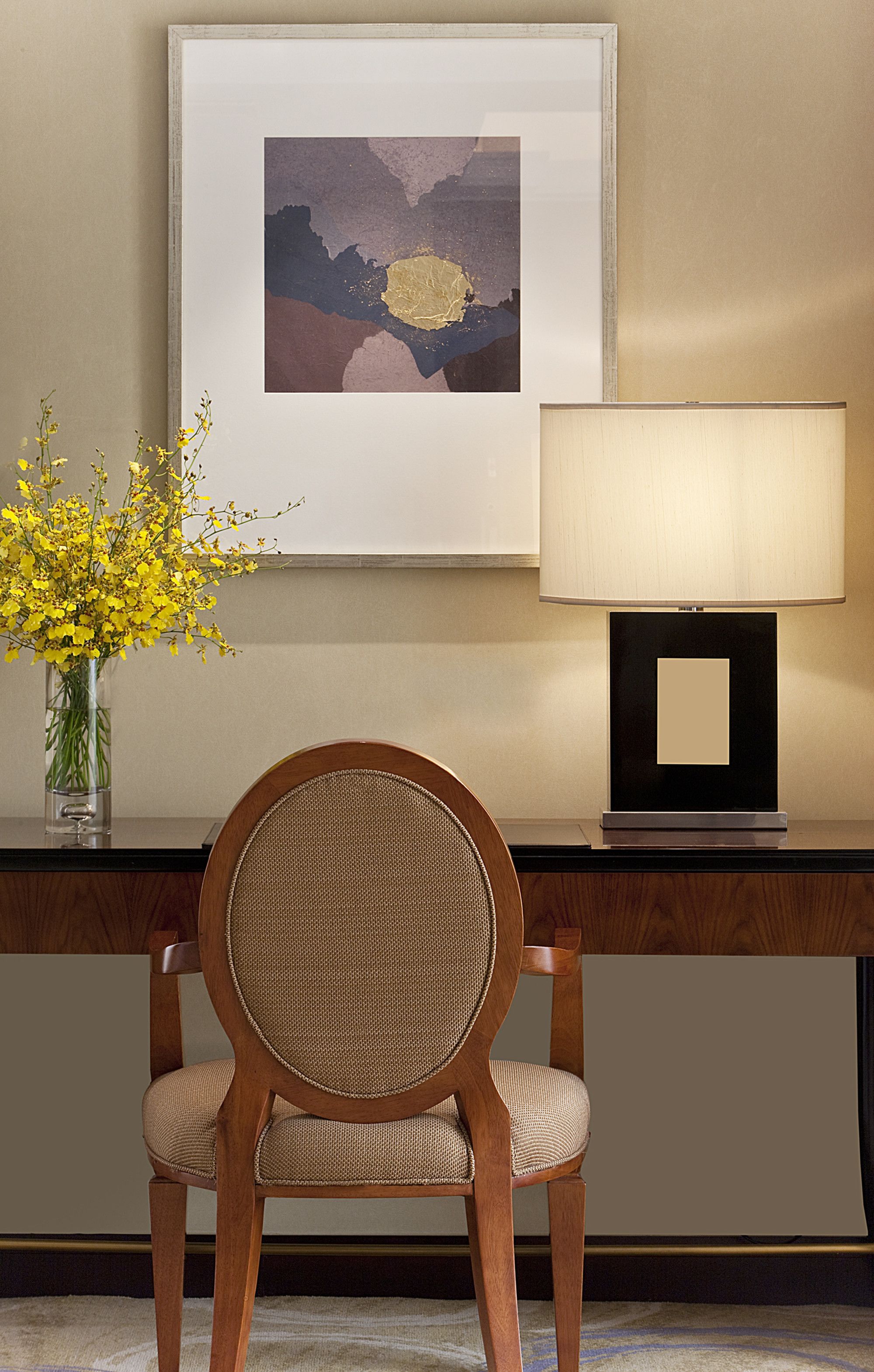 A framed abstract painting, a table lamp, and a vase with yellow flowers on a desk.