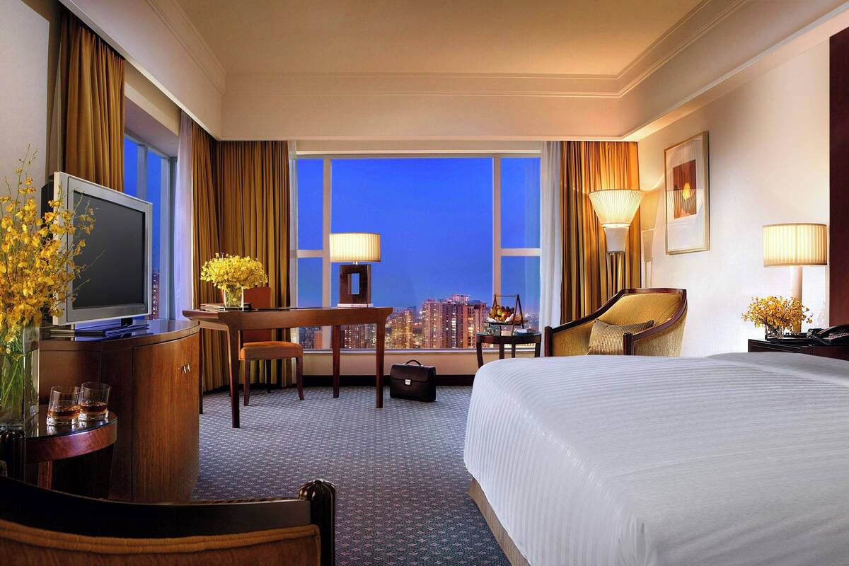 Quarto de hotel com uma cama grande, uma mesa com cadeira, televisão e vista para a cidade ao entardecer.