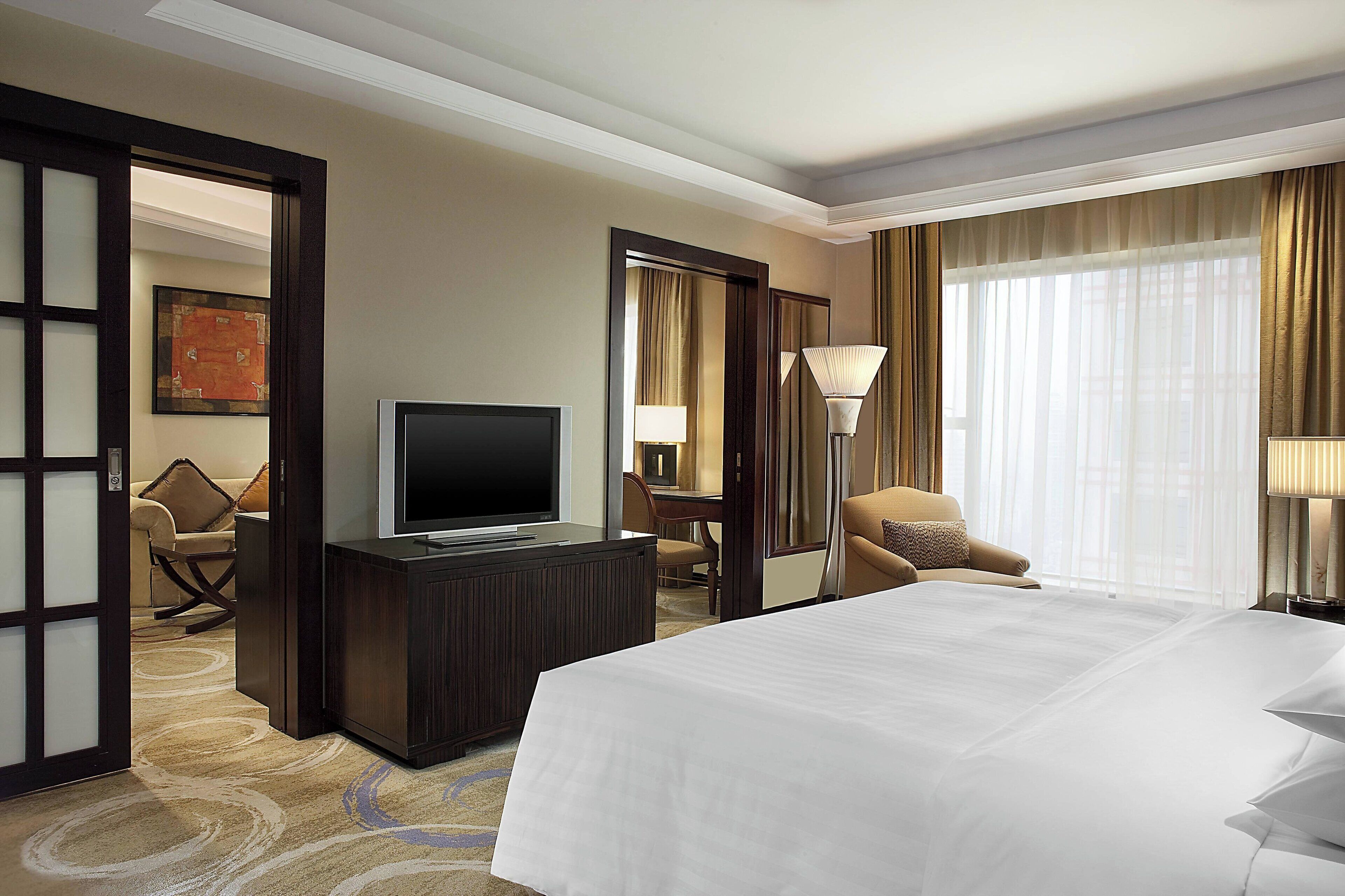 A hotel room with a large bed, a TV, a desk with a chair, and a view of the city through sheer curtains.