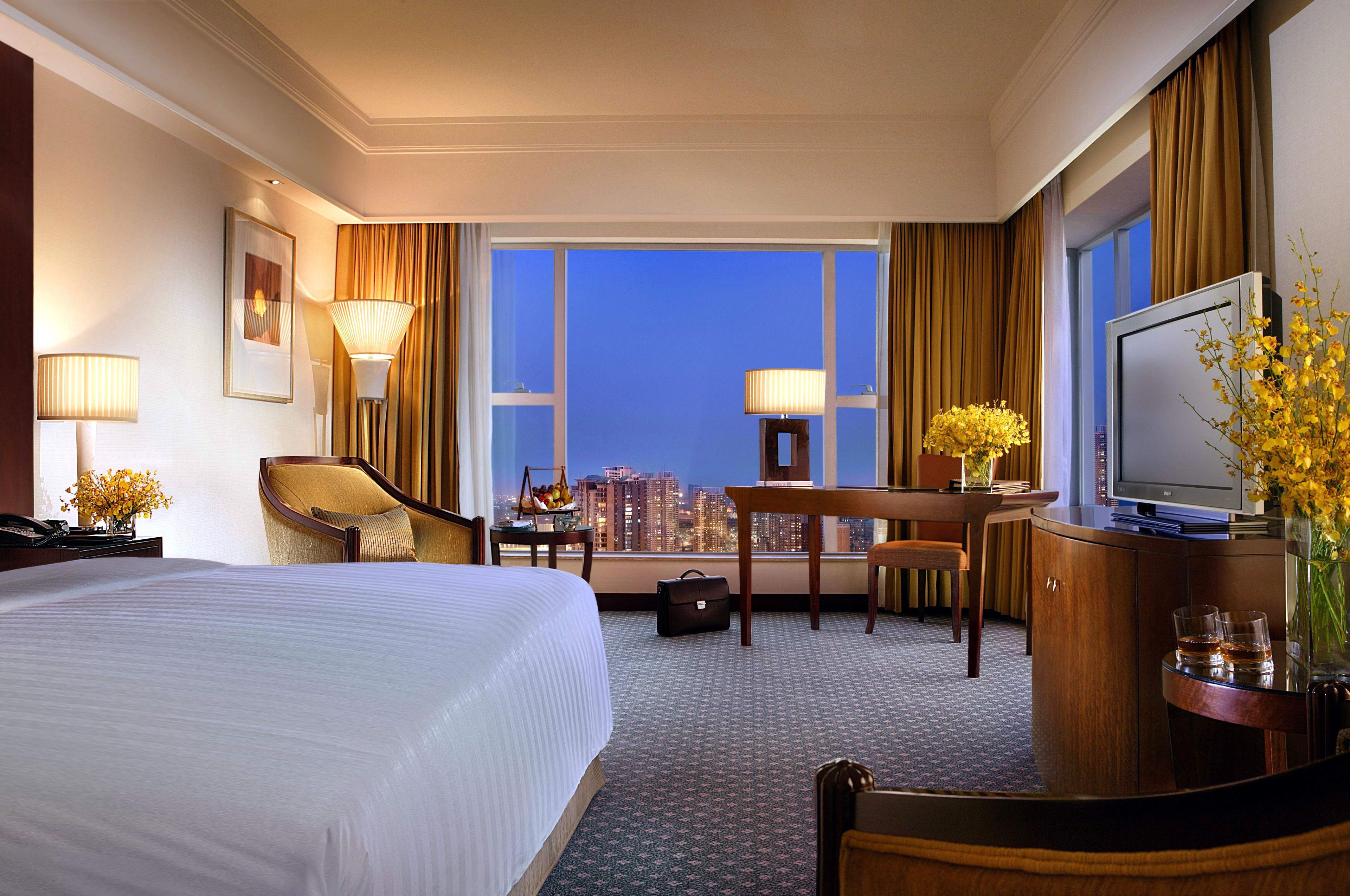A hotel room with a large bed, a desk with a chair, a television, and a view of the cityscape at dusk.