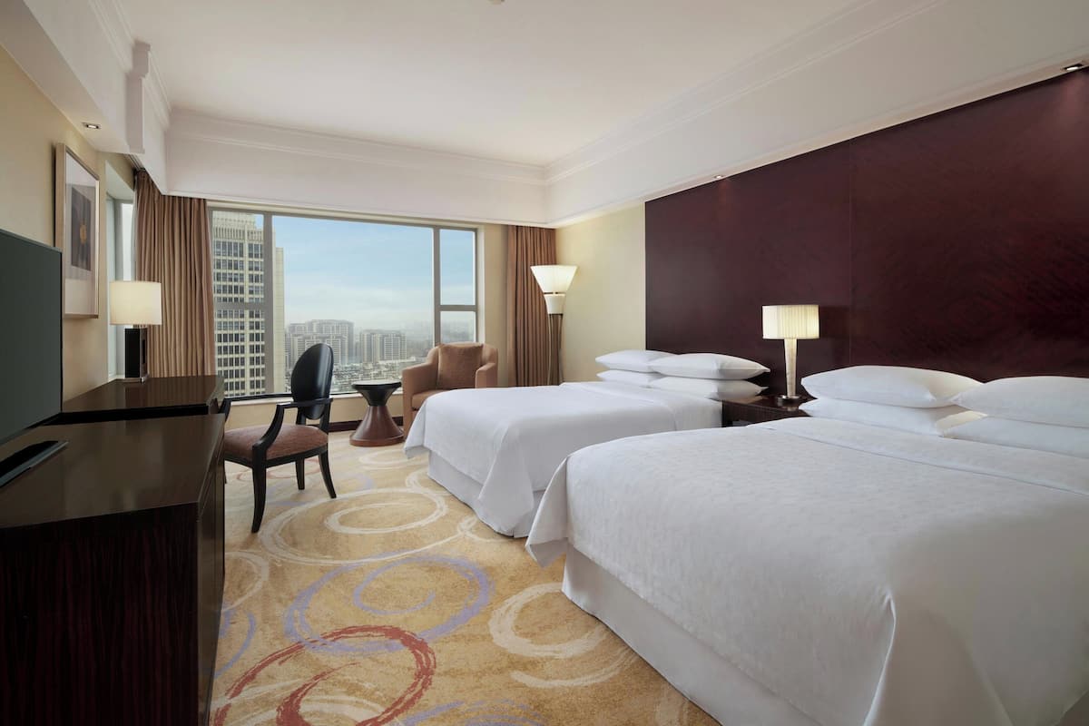 Quarto de hotel com duas camas, uma escrivaninha, uma cadeira e uma janela grande com vista para a cidade.