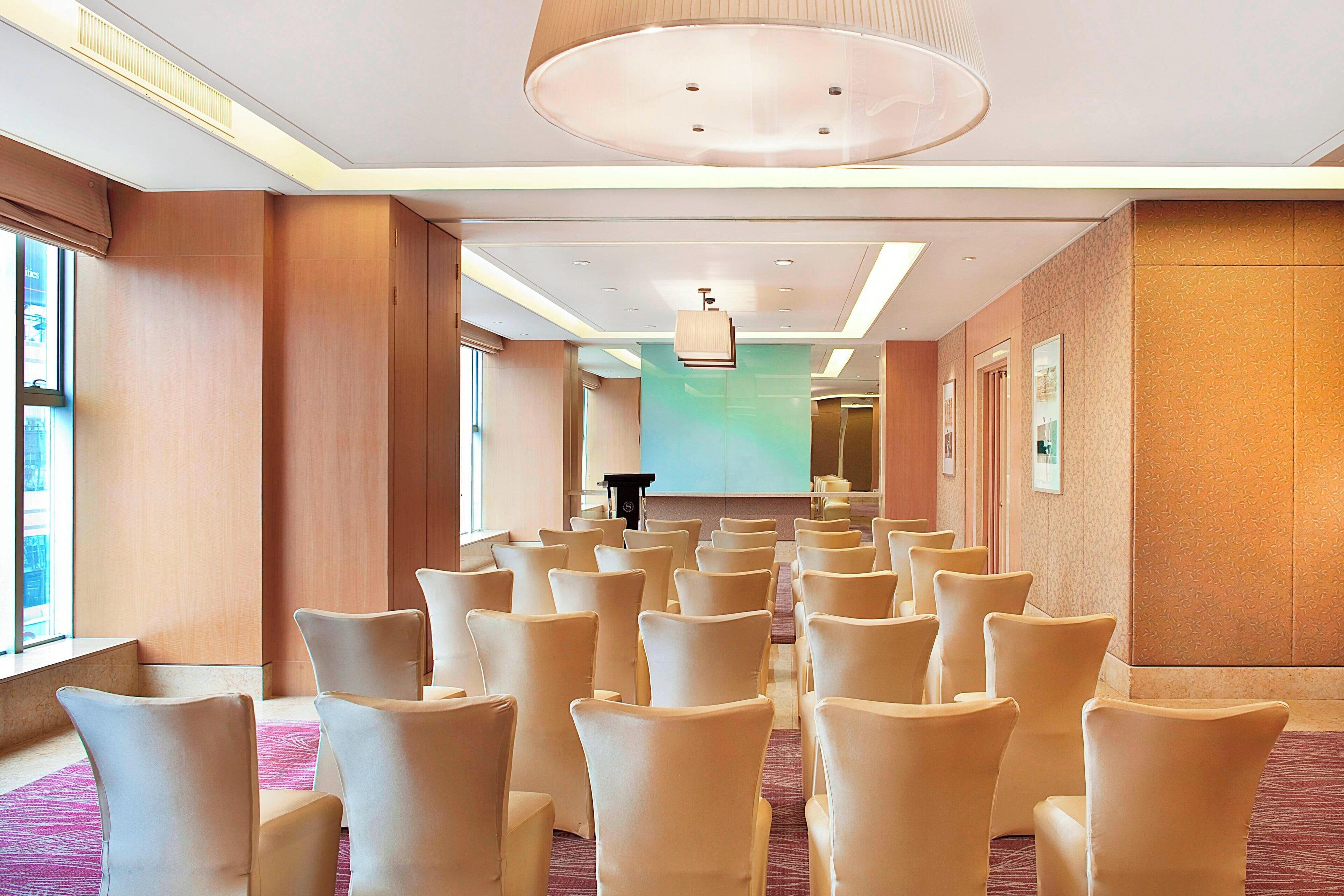 A conference room with rows of chairs facing a projection screen, a podium, and a large window on one side.