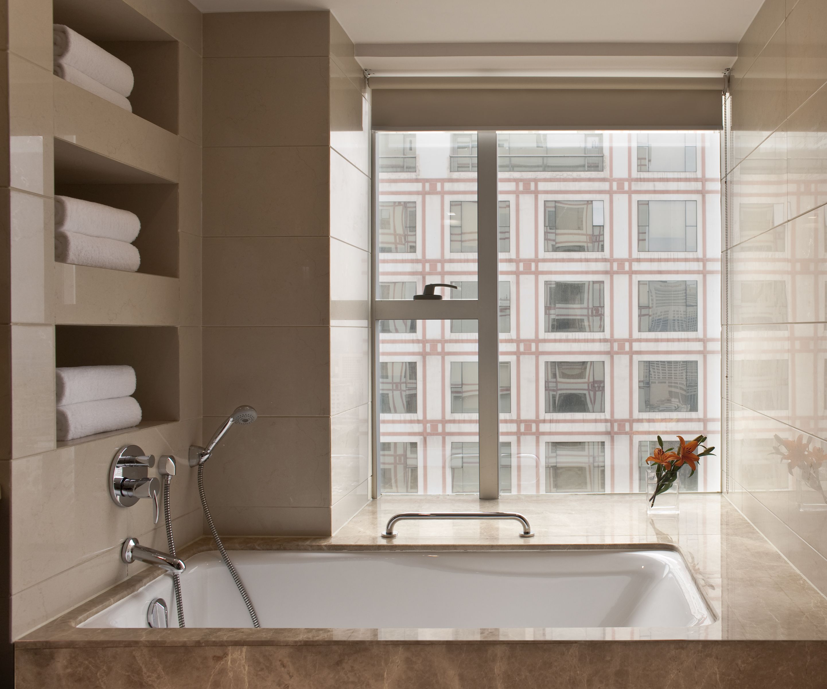 Un baño moderno con bañera, cabina de ducha de vidrio y vistas a un edificio a través del ventanal.