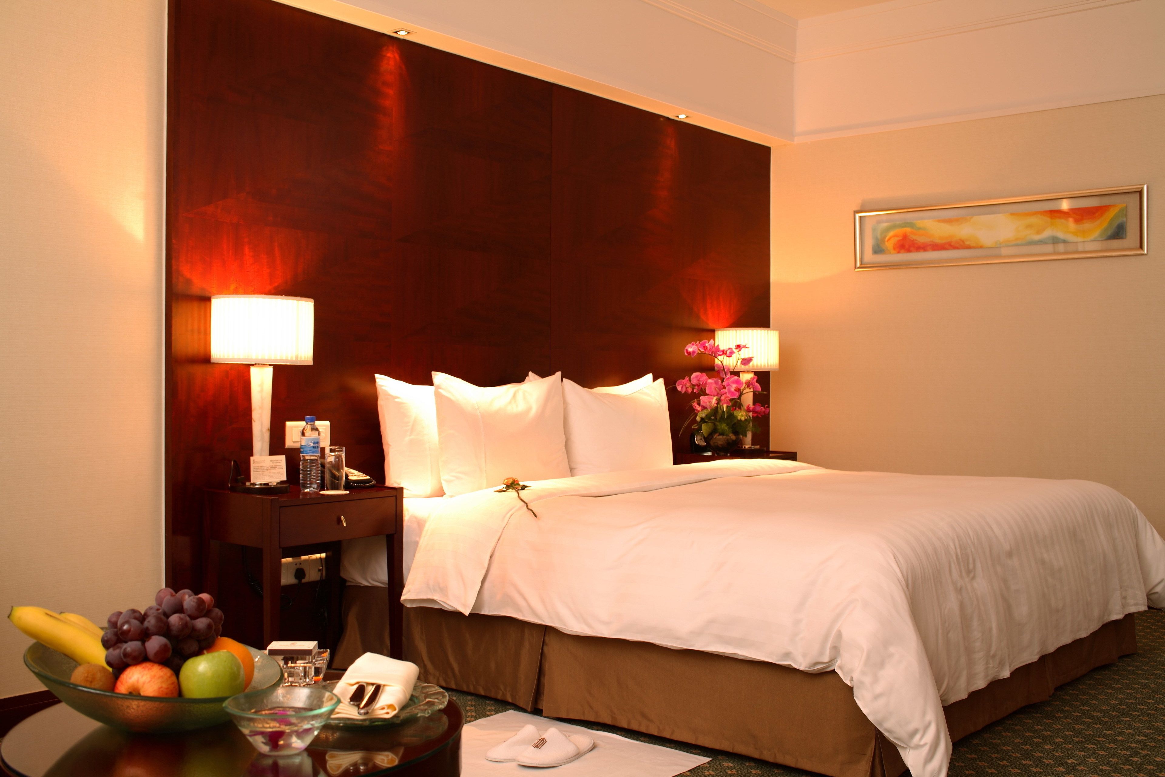 A hotel room with a large bed, two bedside lamps, a nightstand with a phone, a bowl of fruit, and a vase with flowers.