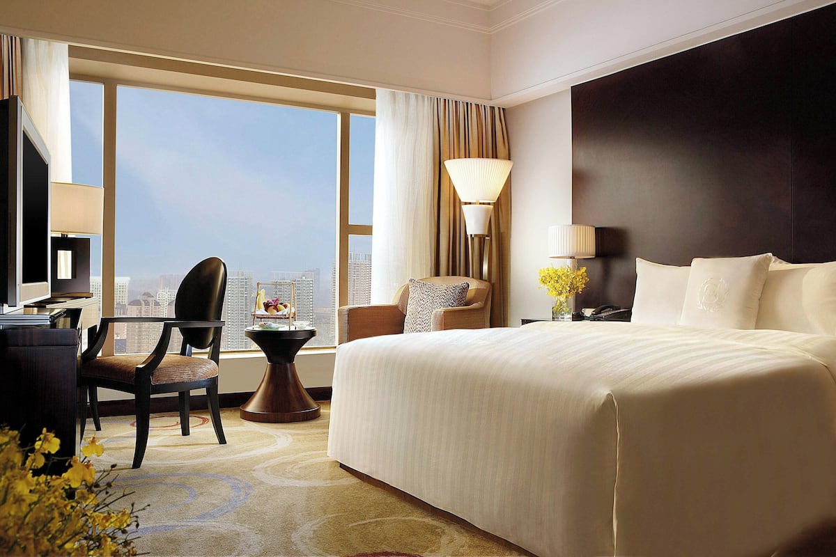 Quarto de hotel com uma cama grande, uma escrivaninha, uma cadeira, um abajur e vista para a cidade.