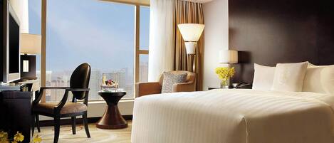 A hotel room with a large bed, a desk, a chair, a lamp, and a view of the city.