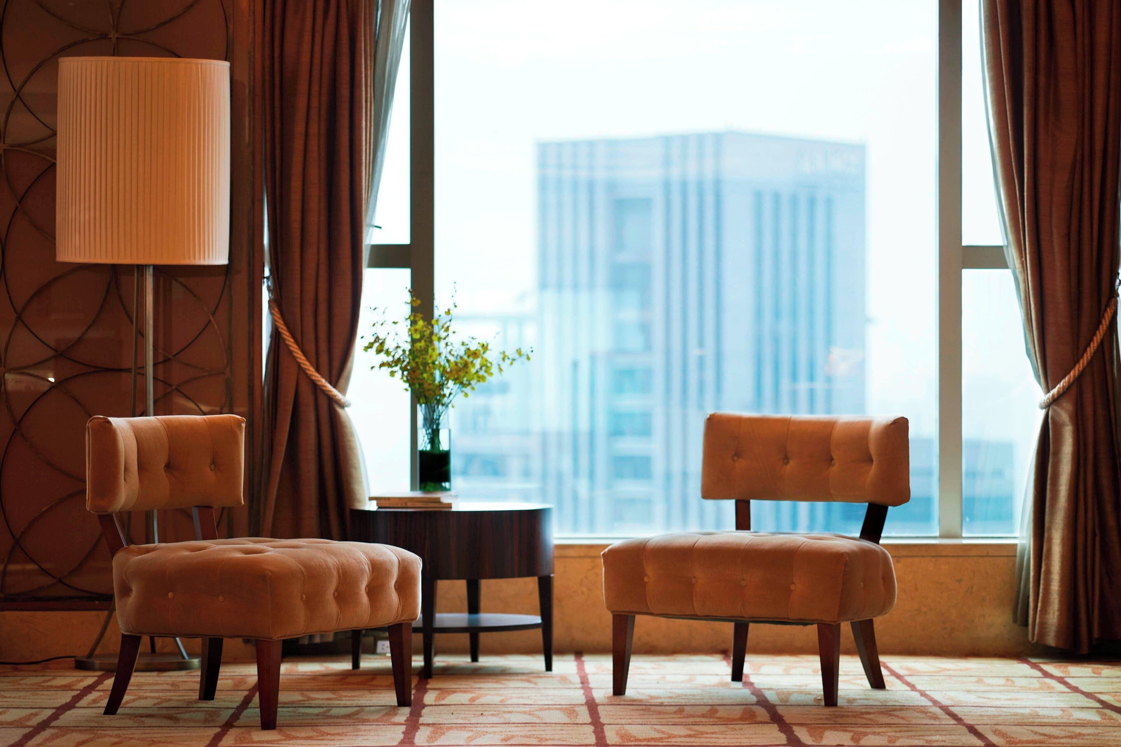 A room with two tufted chairs, a floor lamp, a small table with a vase, and a view of a cityscape through large windows.