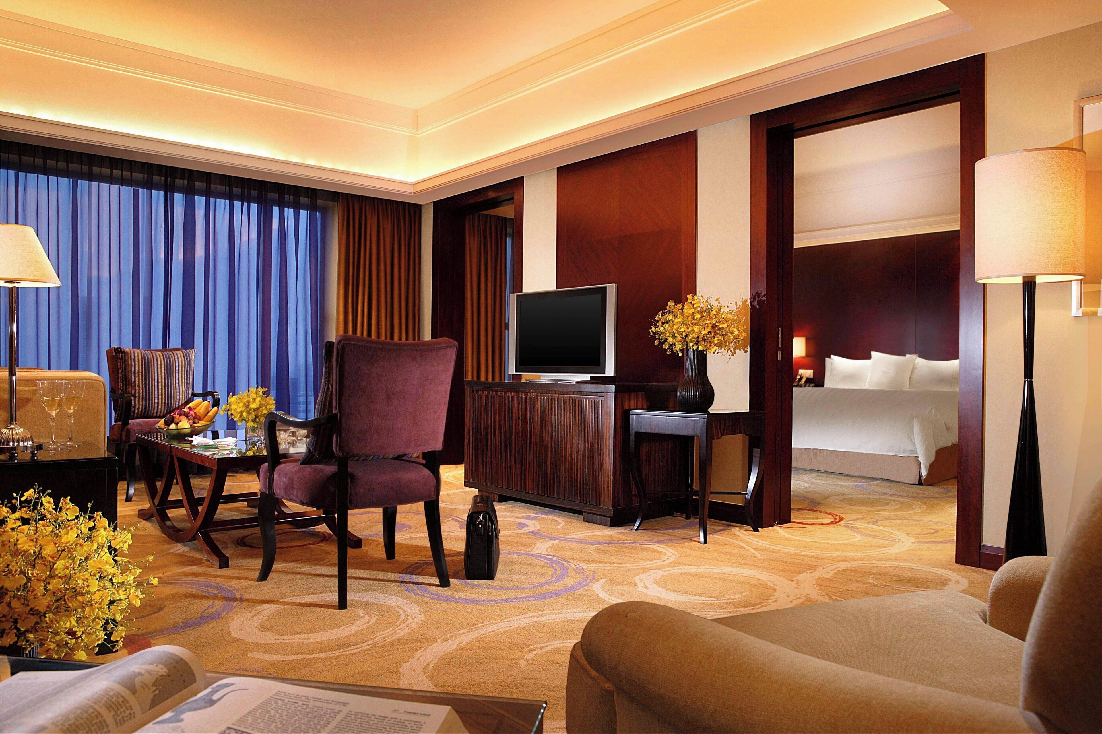 A hotel room with a television, a desk with a computer, a chair, a bed, and a large window with curtains.