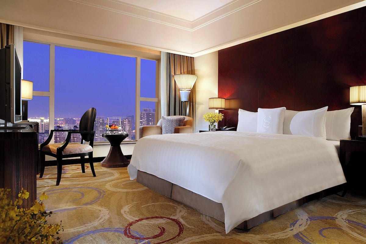 Quarto de hotel com uma cama grande, uma escrivaninha com computador, uma cadeira e vista para a cidade ao entardecer.