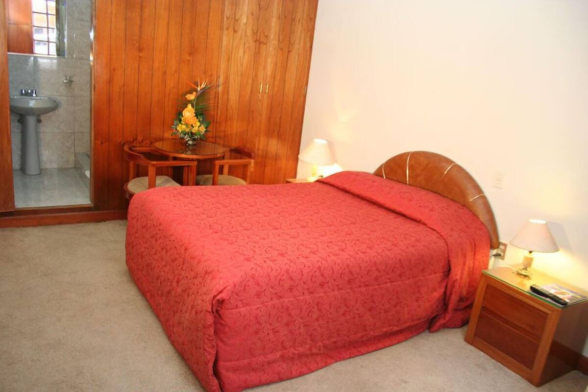 Une chambre à coucher avec une armoire en bois, une table de chevet avec une lampe, une petite table sur laquelle est posé un vase de fleurs et une couverture de lit rouge.
