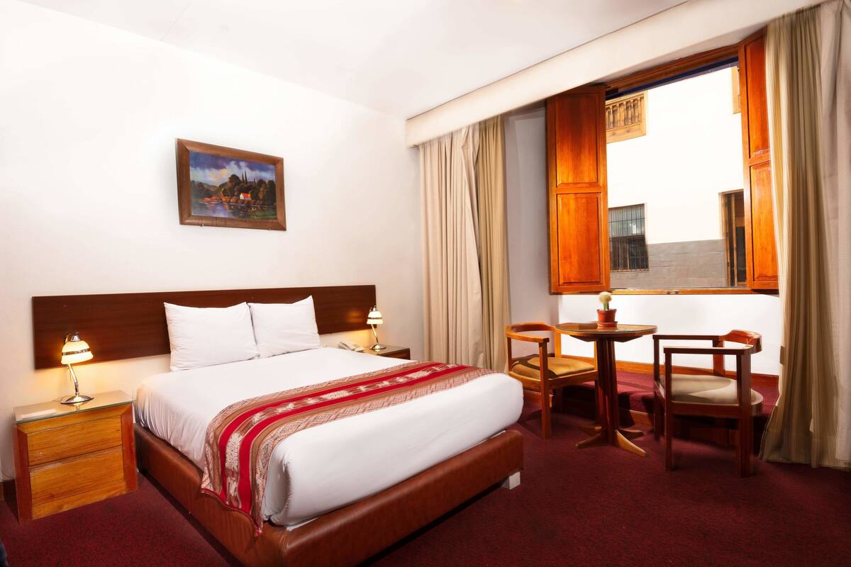 Une chambre d’hôtel avec un grand lit, un bureau, deux chaises, une table de chevet avec une lampe, un tableau au mur et une fenêtre avec des rideaux.