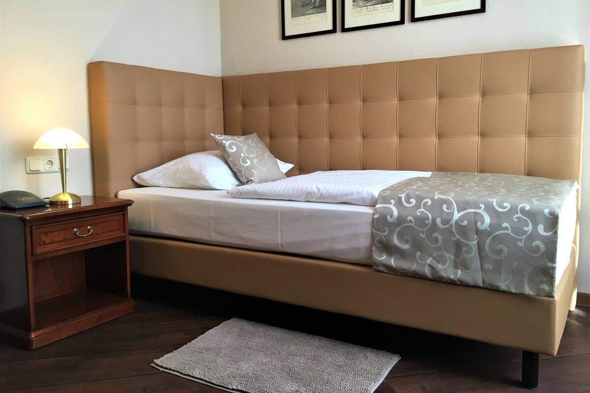 Una cama bien tendida con un cabecero color beige, una mesita de noche con lámpara, y una alfombra en el suelo.