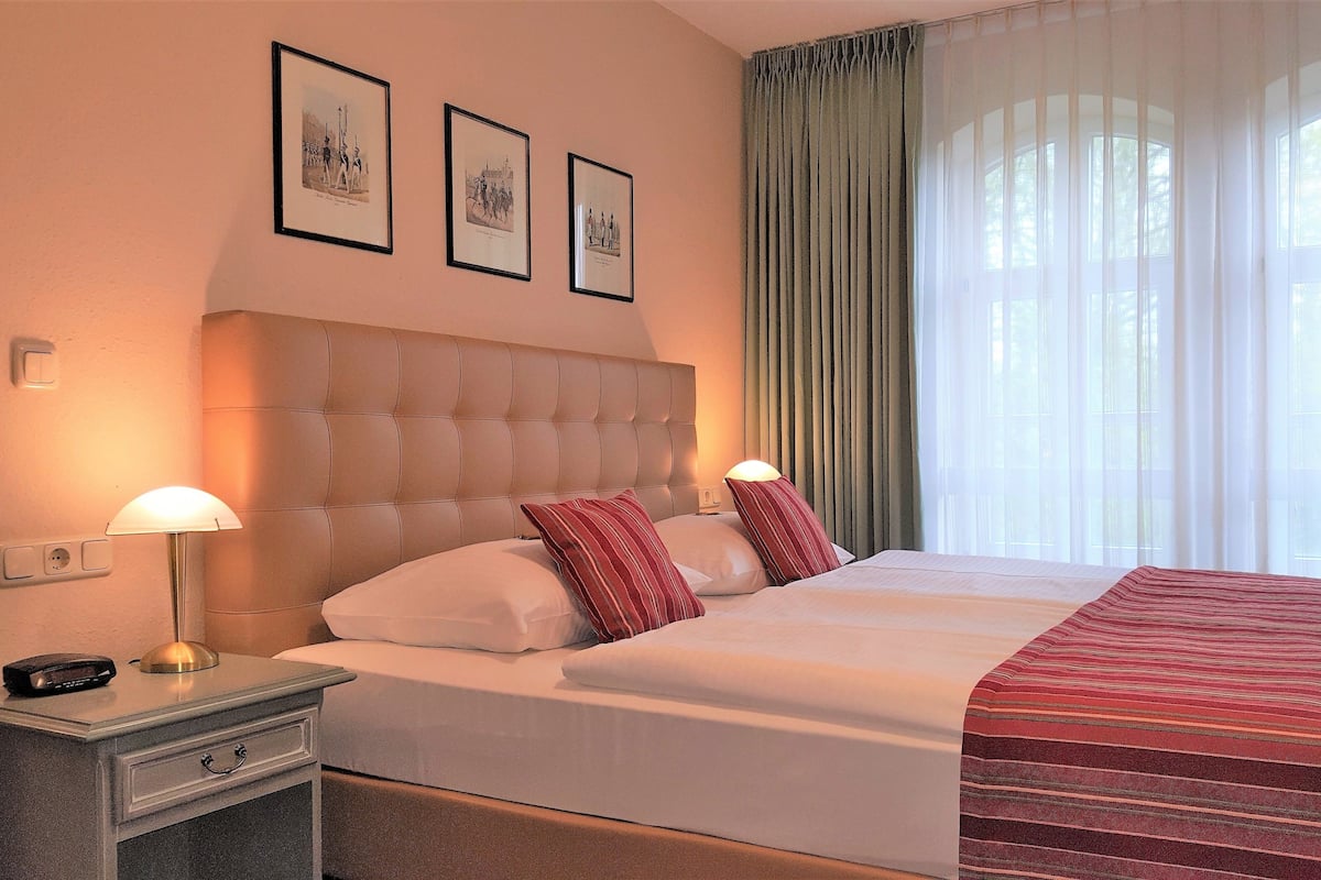 Una habitación de hotel con una cama grande, dos lámparas de noche, una mesita de noche y un ventanal con cortinas traslúcidas.
