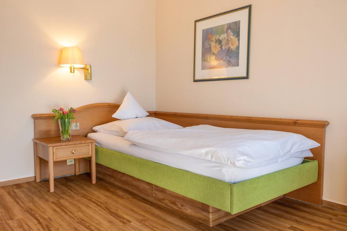 A neatly made bed with white linens, a bedside table with a lamp, and a framed floral painting on the wall.