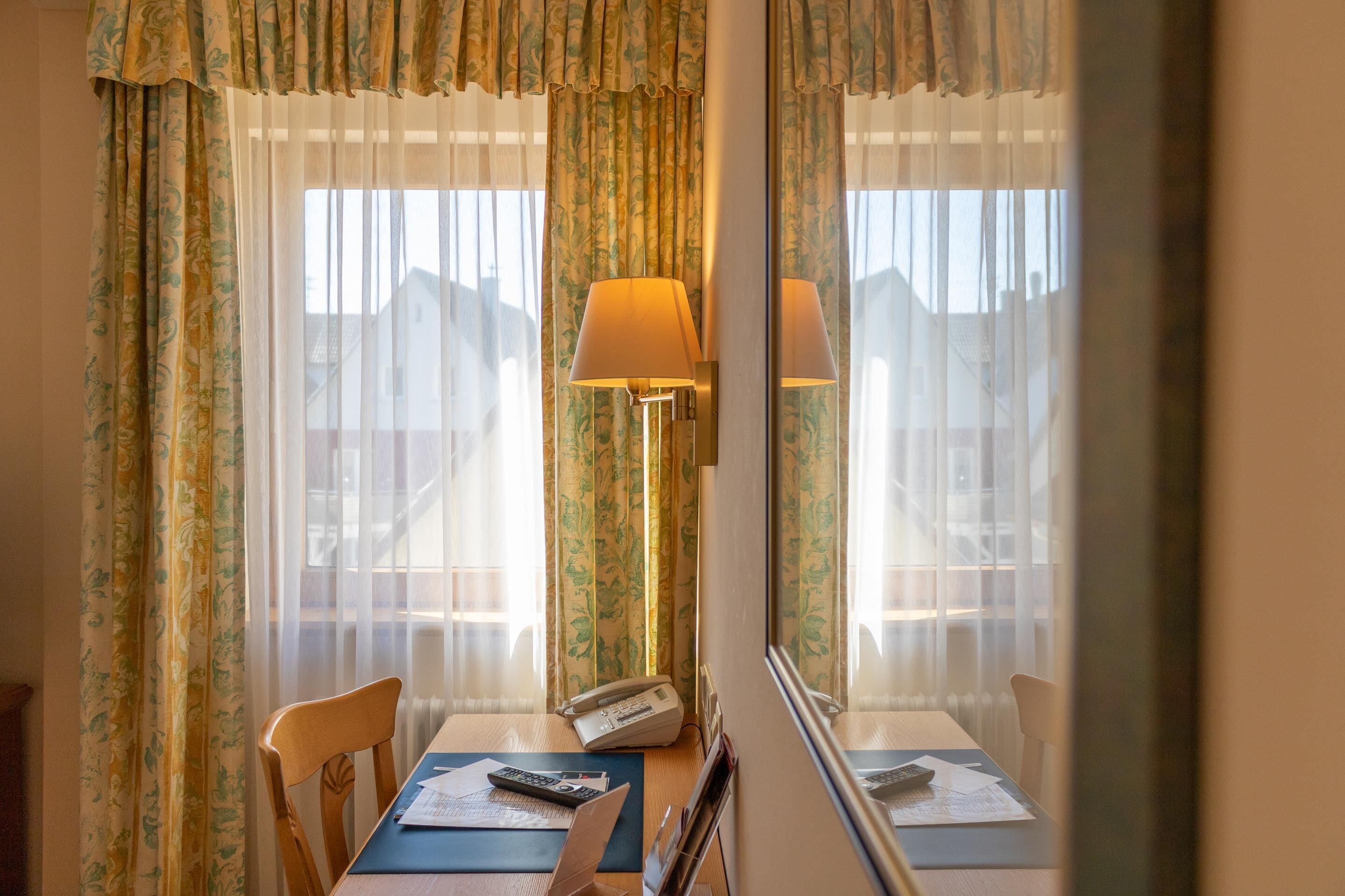 A hotel room with a dining table, chairs, a lamp, a telephone, and a window with curtains.