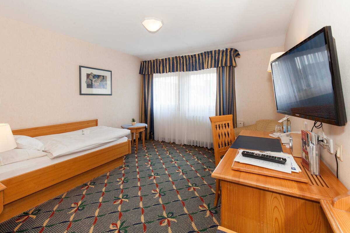 A hotel room with a bed, a desk with a TV, a chair, a small table, and a window with curtains.
