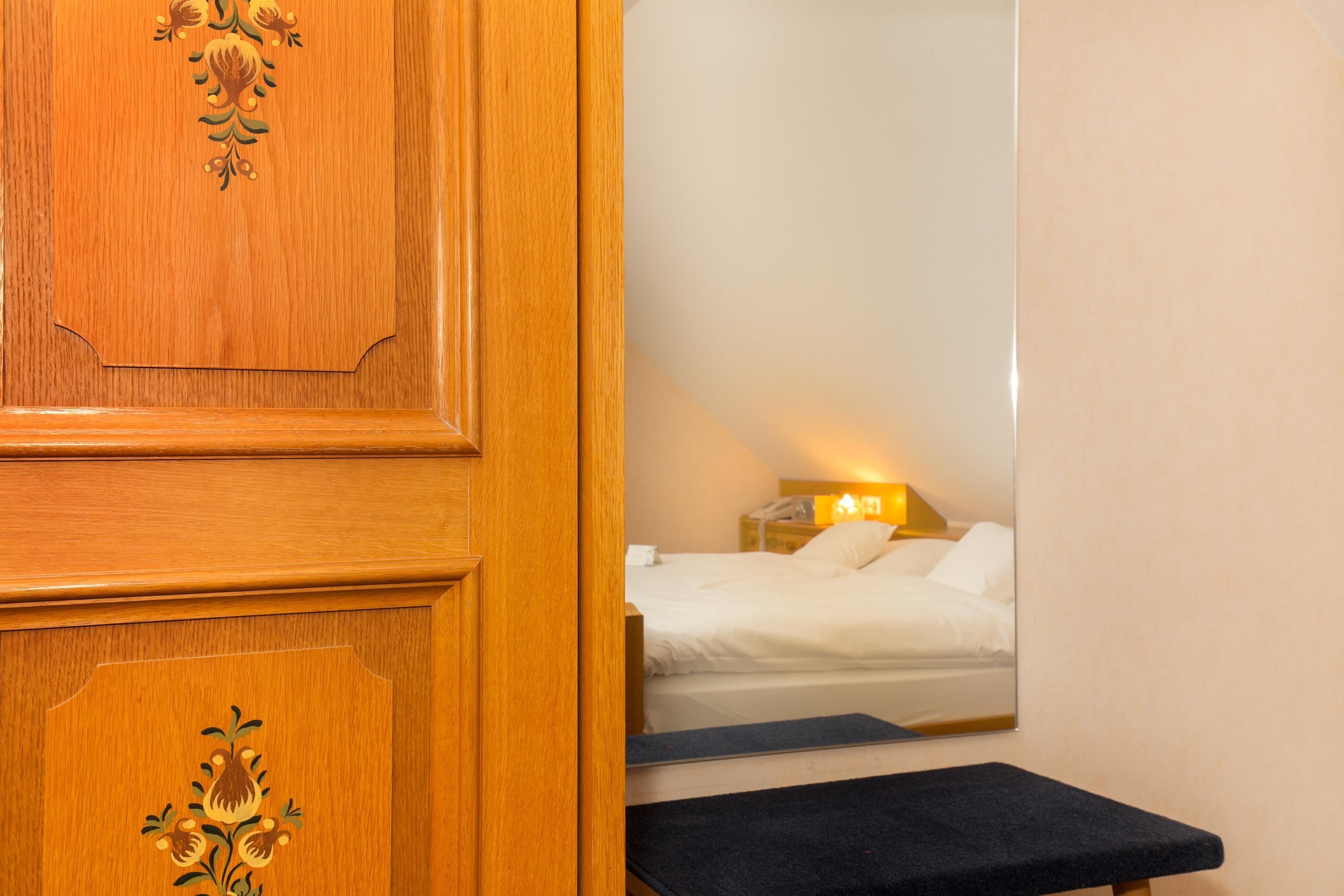 Una puerta de madera con adornos florales, una cama con sábanas blancas, una lámpara de noche y un pequeño banco.
