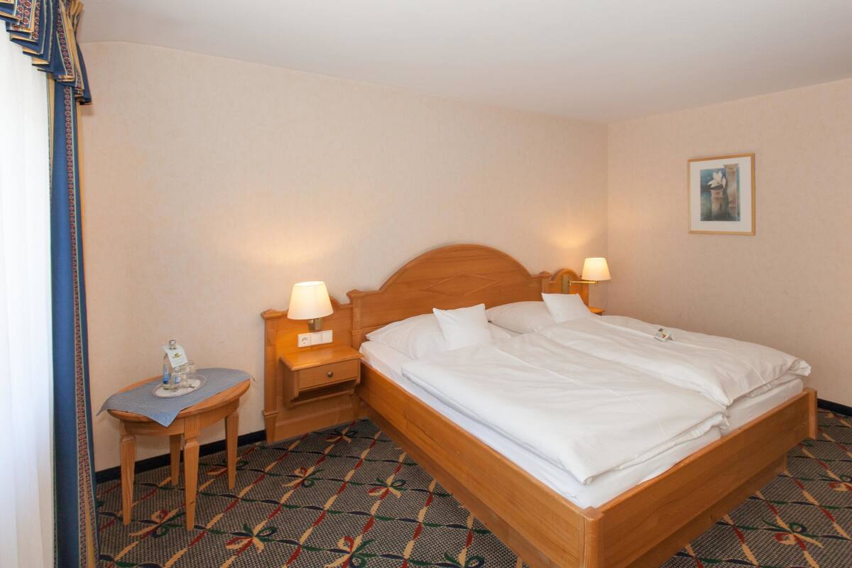 A hotel room with a wooden bed, two bedside tables with lamps, a small table with a placemat, and a patterned carpet.