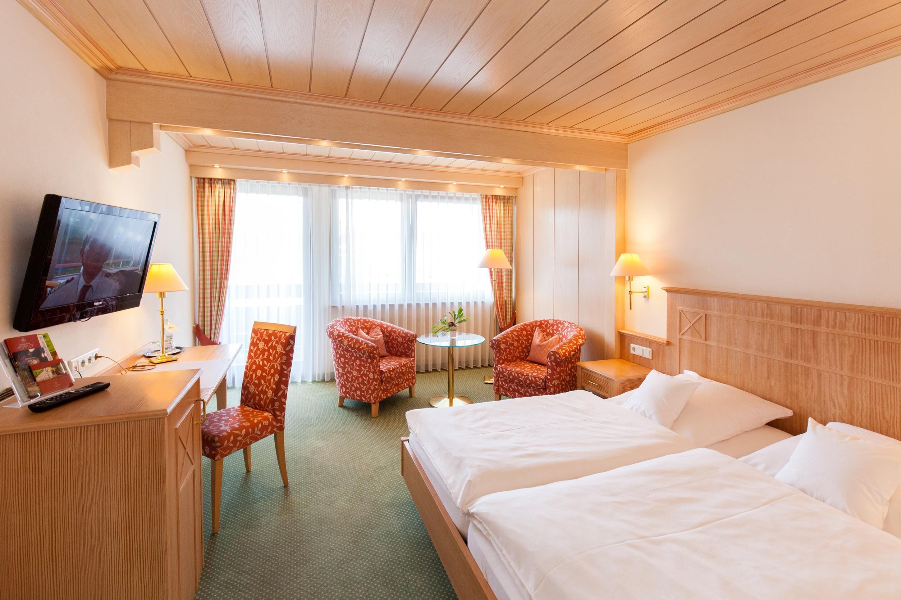A hotel room with a bed, two armchairs, a desk with a TV, lamps, and a window with curtains.